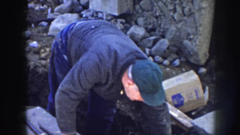 The man bends low beside a plank and marked cardboard box at the Nebraska railway worksite, with rocks and dirt surrounding the trench.