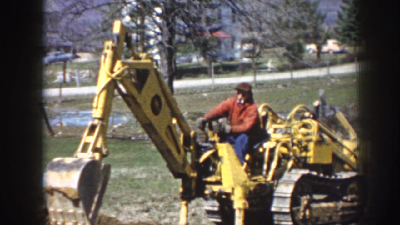 Adult man in a red jacket operates a yellow tracked excavator in a grassy Nebraska setting with trees and houses behind him.