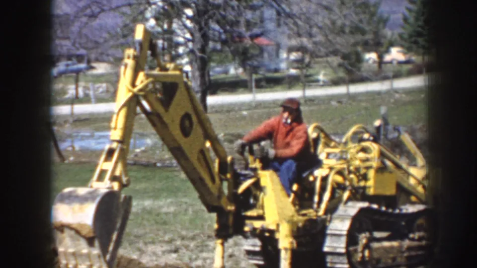 Adult man in a red jacket operates a yellow tracked excavator in a grassy Nebraska setting with trees and houses behind him.
