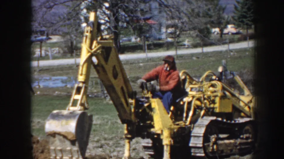 The excavator operator is seen from farther back, with the bucket arm raised over a grassy area in Nebraska 1958 footage.