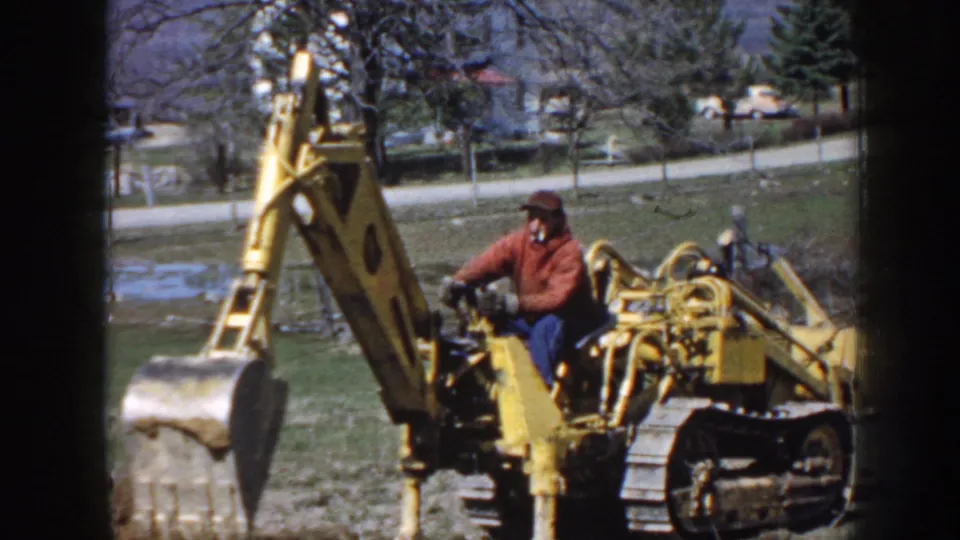 A man in a red jacket operates a yellow tracked excavator near grass, trees, and distant houses in archival Nebraska footage from 1958.