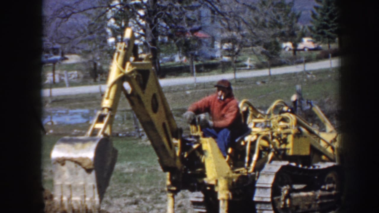 Man in red jacket operates a yellow tracked excavator in a grassy field.