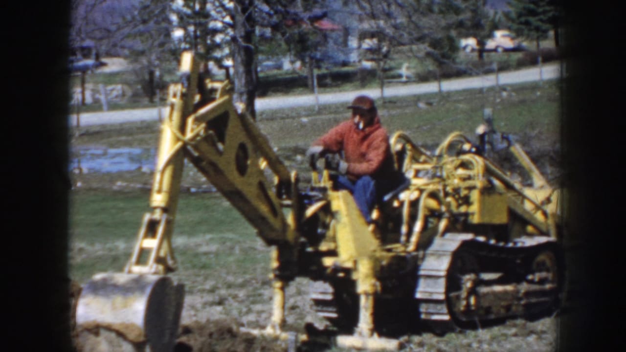The 1958 Nebraska frame shows the operator seated on a yellow tracked excavator, with the digging arm raised near a rural roadside.