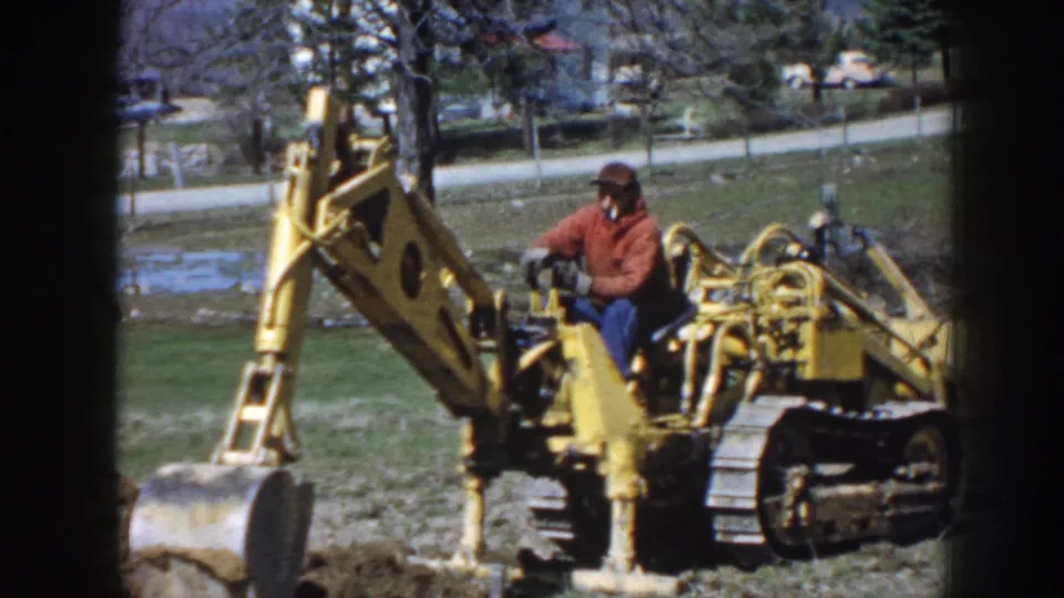 The 1958 Nebraska frame shows the operator seated on a yellow tracked excavator, with the digging arm raised near a rural roadside.
