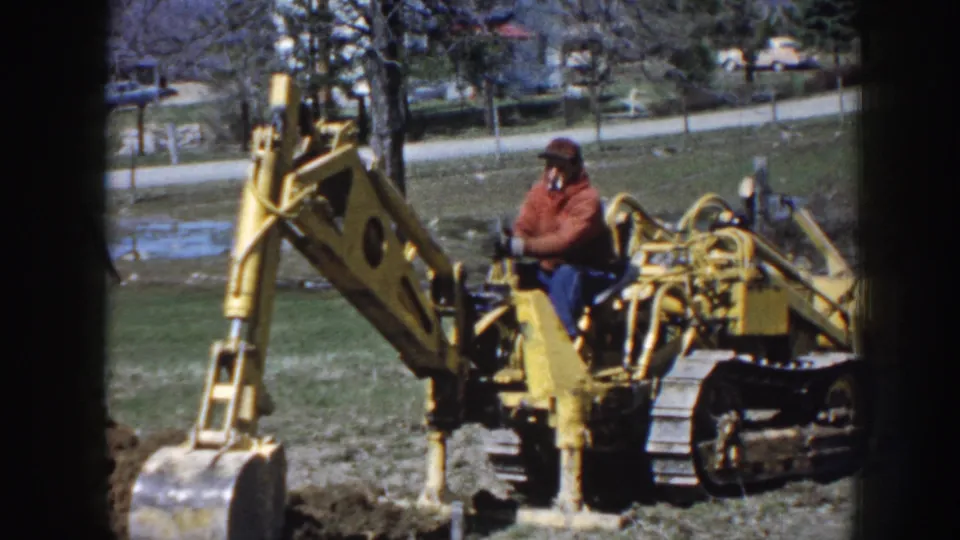 A small yellow tracked excavator is shown digging in a Nebraska field, with the operator in a red jacket seated at the controls.
