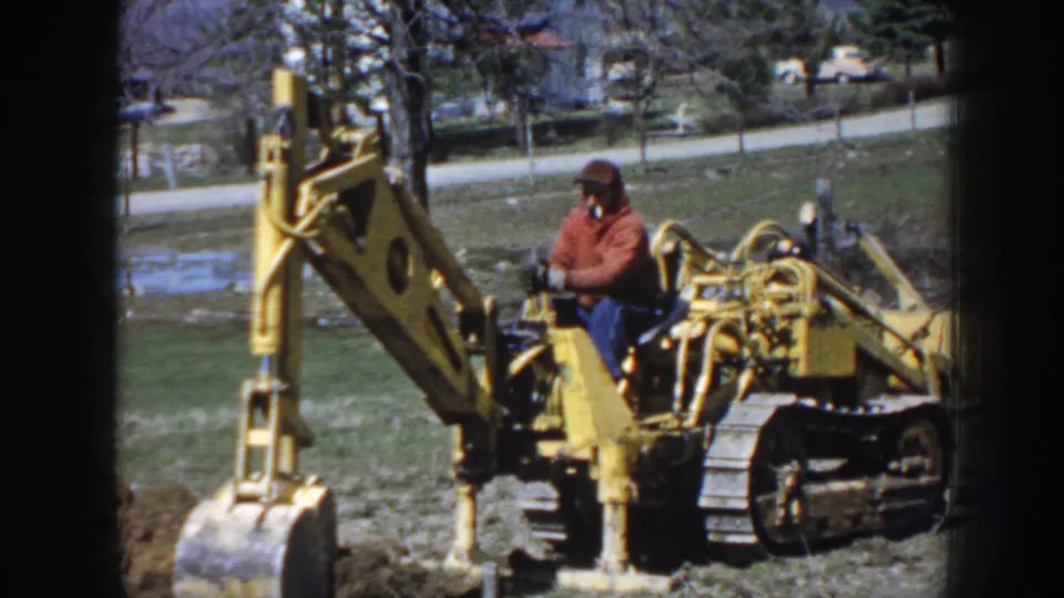 The final frame shows the man on the yellow excavator in the open Nebraska field, with road, trees, and parked cars beyond.