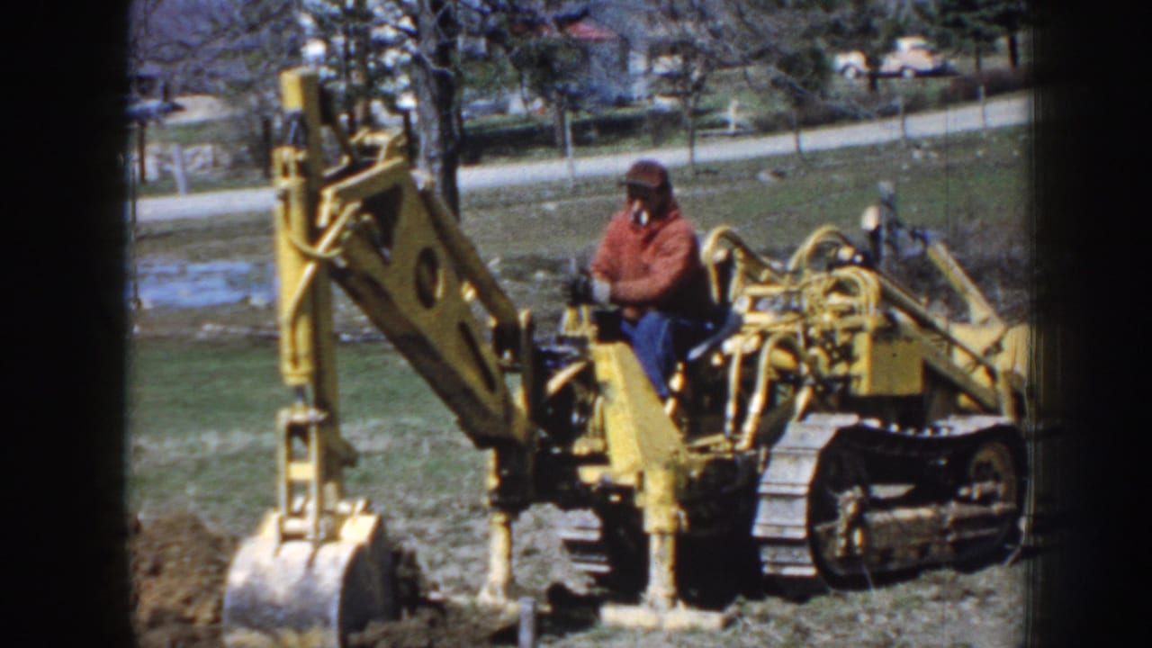 Man in a red jacket operating a yellow tracked digger in a grassy yard.
