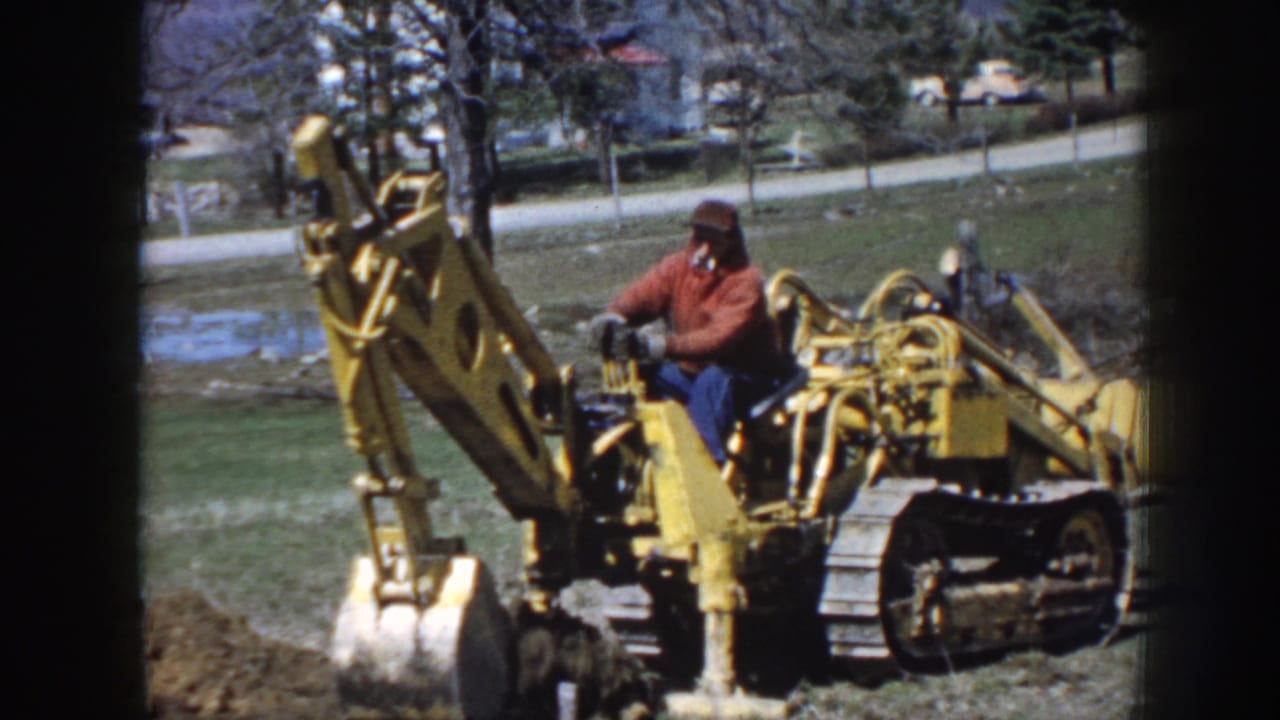 1958 Nebraska home-movie frame with a man seated on a yellow tracked digger as its bucket works near exposed earth.