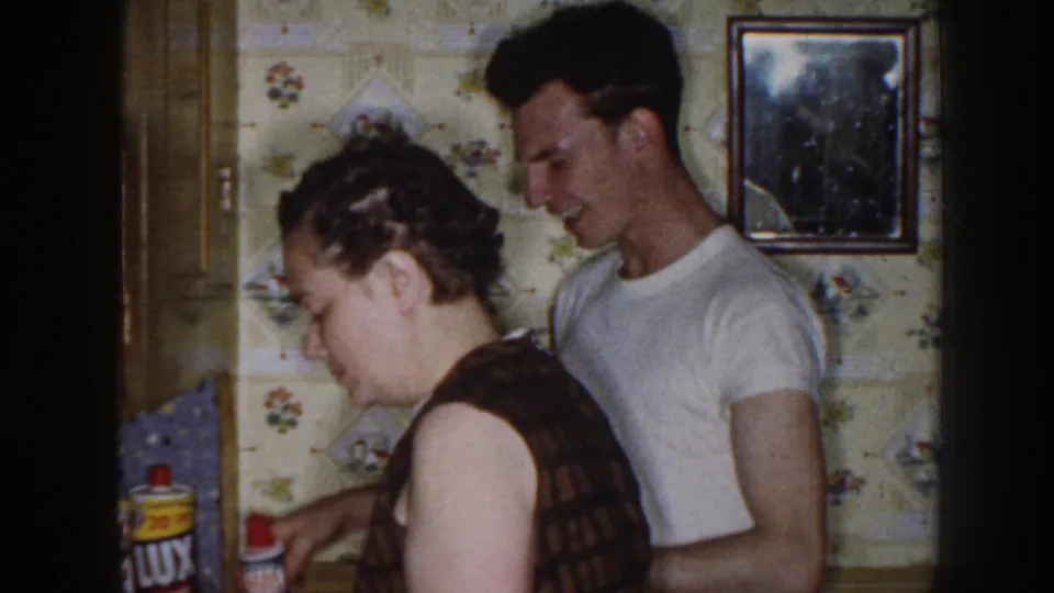 A woman in a dark sleeveless dress and a man in a white T-shirt stand in a Nebraska kitchen with floral wallpaper.