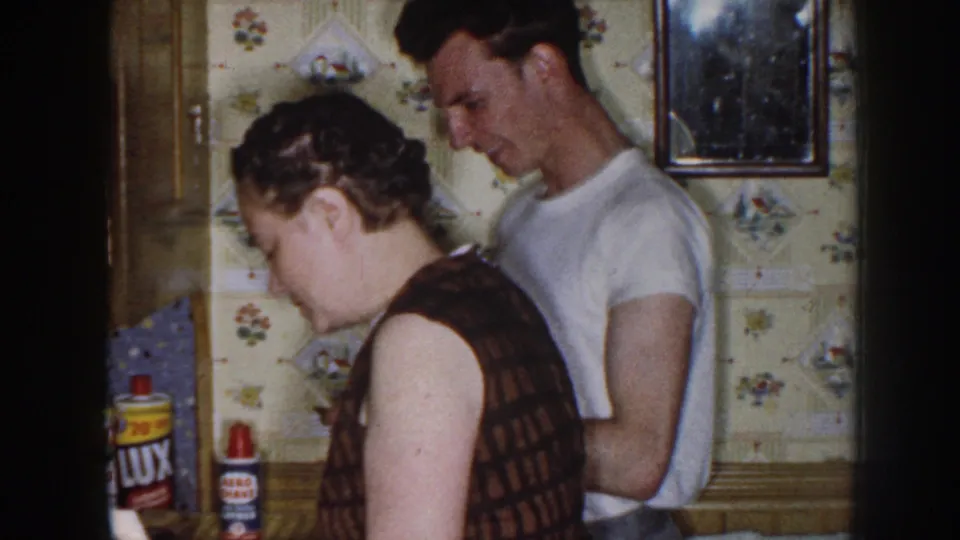 The couple remains in the kitchen, with the woman turned toward the counter and product packaging visible nearby.