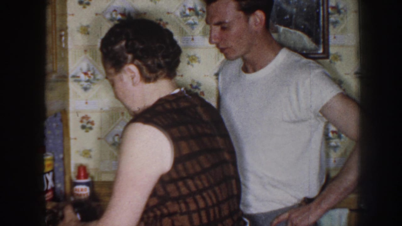 1958 Nebraska home-movie frame with a woman at the counter and a man standing nearby, with household product labels visible in the kitchen.