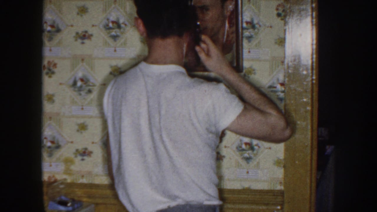Young man in a white t-shirt stands with his back to the camera at a wall mirror.