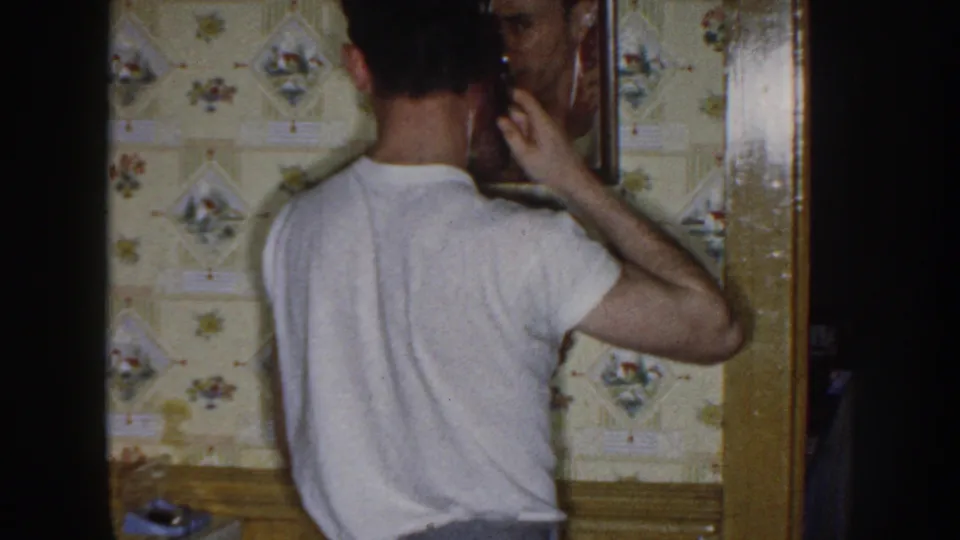 In a 1958 Nebraska home interior, a young man in a white t-shirt faces a wall mirror beside floral wallpaper and wood trim.