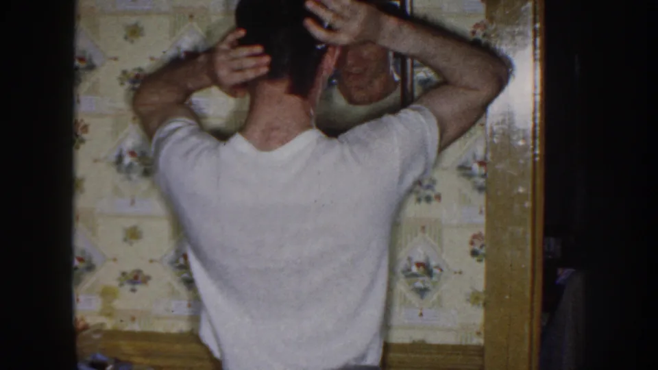 The young man adjusts his hair with both hands in front of a mirror, with floral wallpaper and wood trim visible around him.