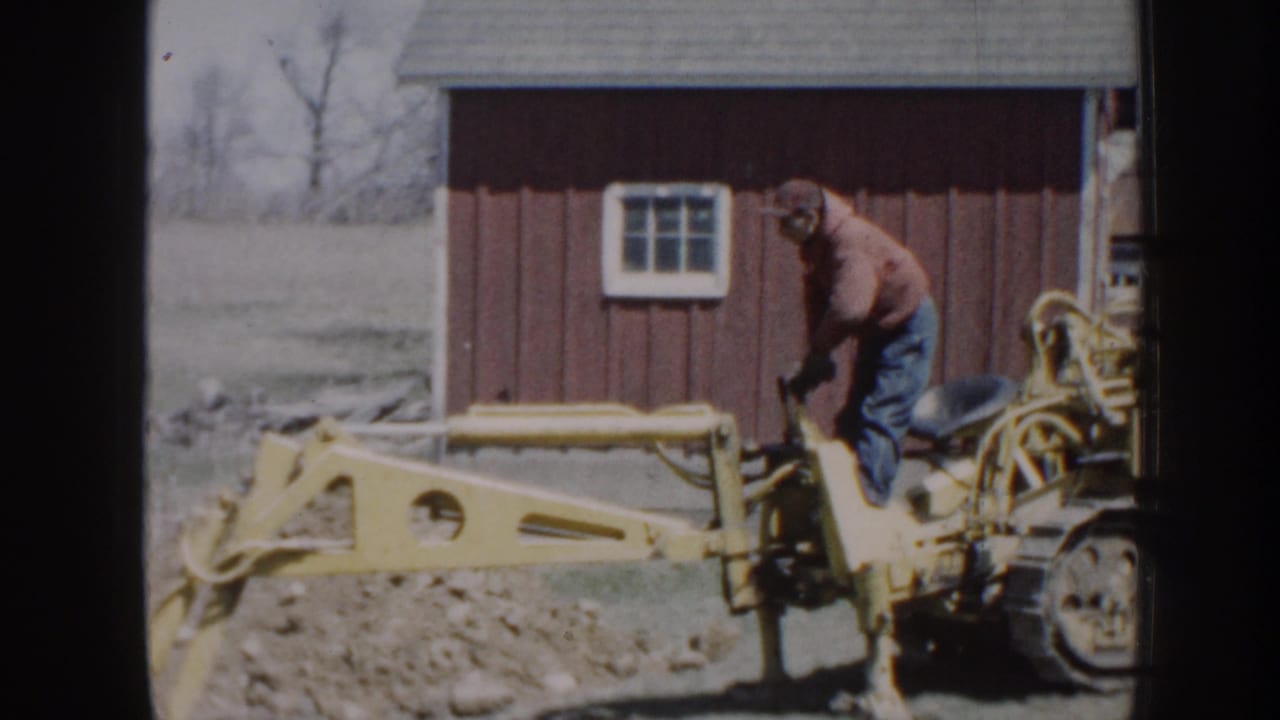 Man in a red hooded top operates a yellow backhoe beside a red outbuilding.
