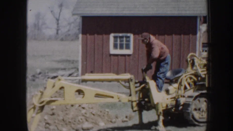 In 1958 Nebraska 8mm footage, a man in work clothes uses a yellow backhoe near dirt piles and a red rural outbuilding.