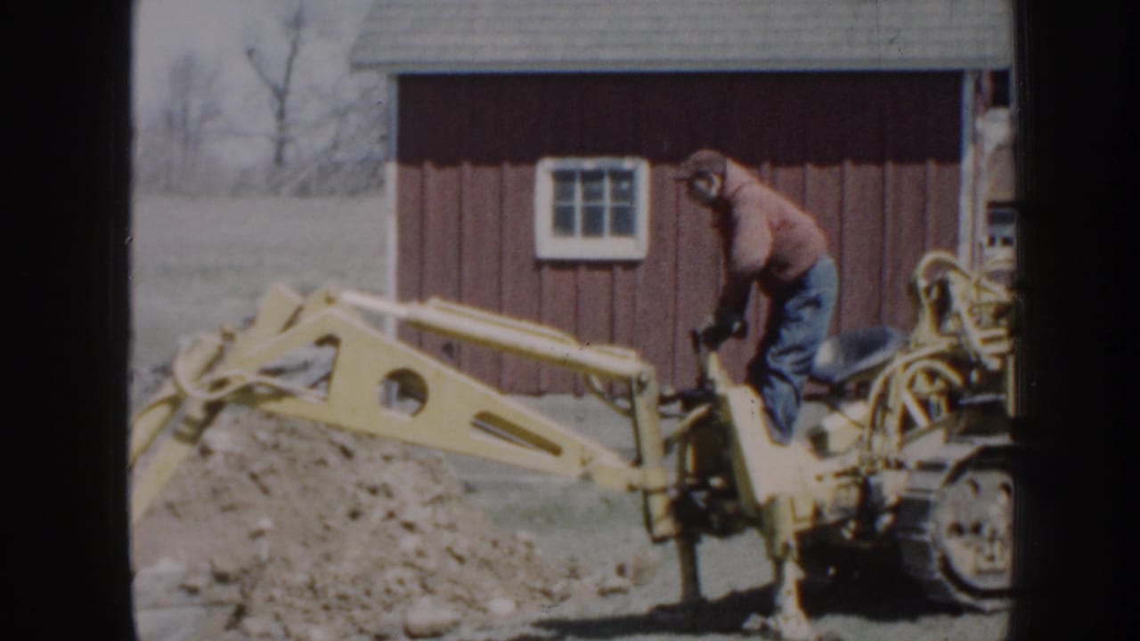 The operator stands on the yellow backhoe while the arm reaches over a dirt pile beside the red outbuilding.