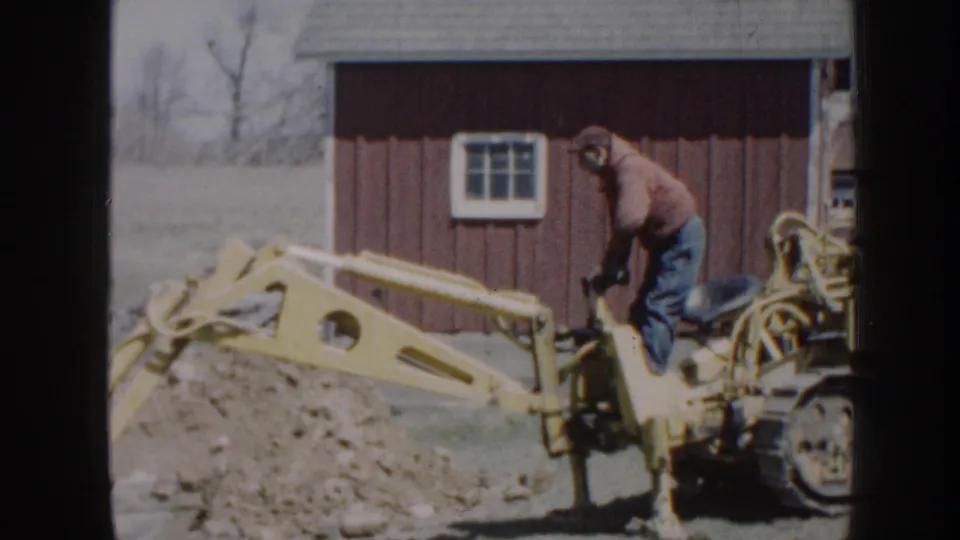 The operator stands on the yellow backhoe while the arm reaches over a dirt pile beside the red outbuilding.