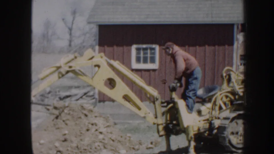 A yellow backhoe bucket lifts near a mound of dirt while the man stands at the controls in a rural Nebraska yard.