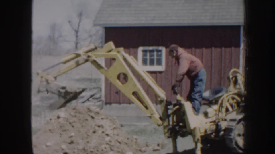 The raised yellow backhoe arm fills the foreground while the man operates the machine near the red outbuilding.