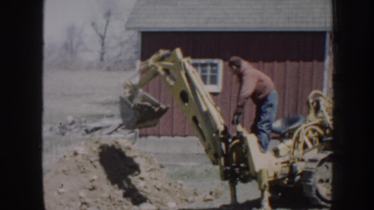 Man operating a yellow backhoe beside a red barn as the bucket digs into soil.