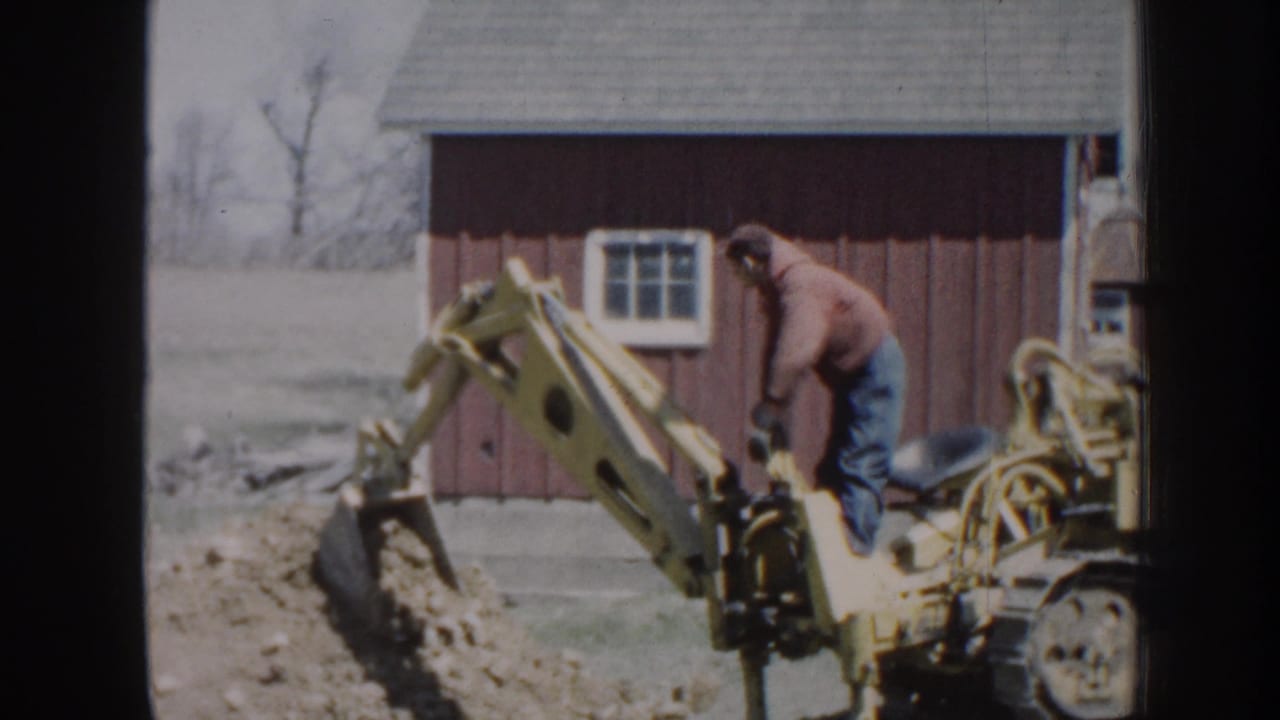 The operator leans over the controls as the yellow backhoe bucket moves dirt beside the red barn on rural Nebraska property.