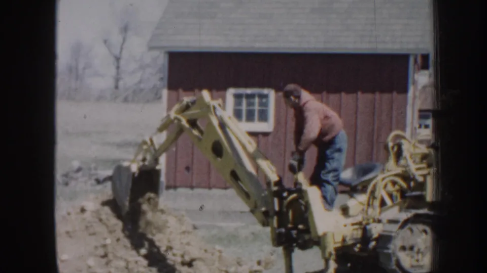 A yellow backhoe continues trenching soil near a red barn, with the adult operator visible in red jacket and jeans.