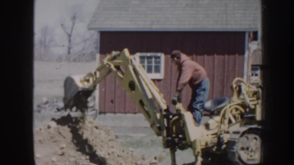 Near the end of the clip, the backhoe bucket lifts dirt beside the red barn while the operator remains at the controls.