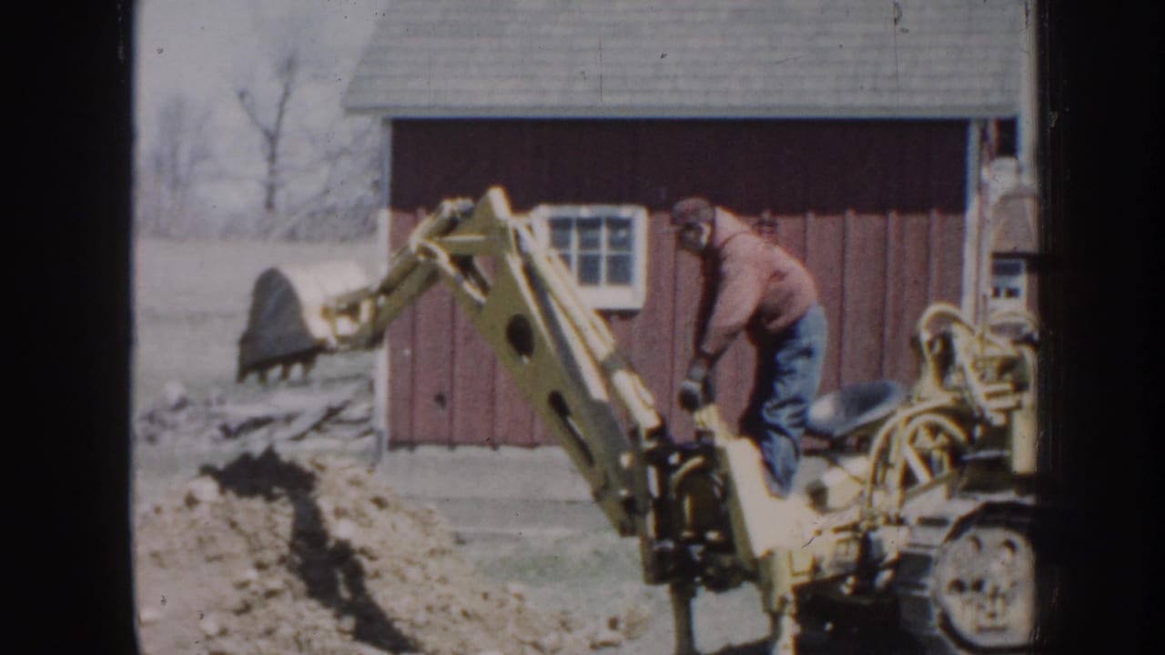 Man operating a yellow backhoe beside a red barn while digging a trench.