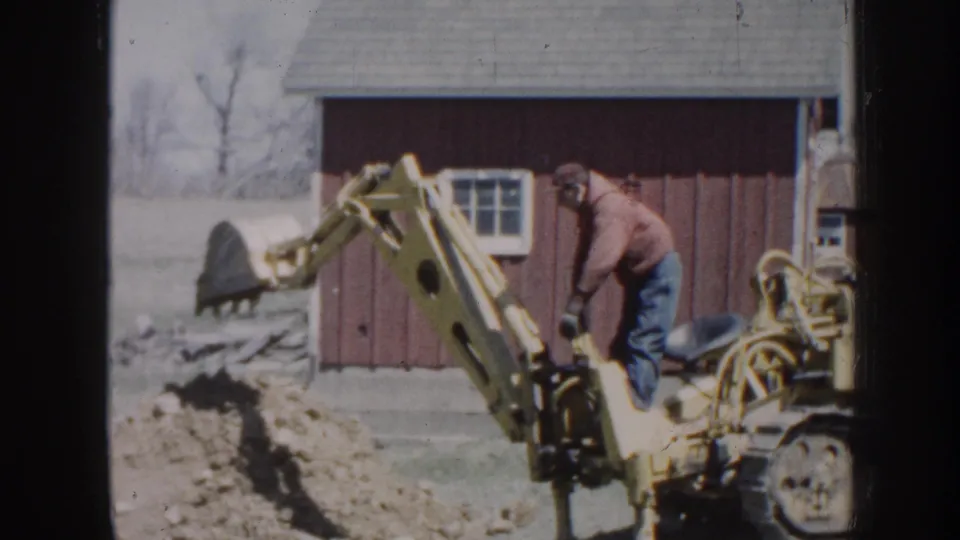 1958 Nebraska home-movie frame showing a man on a yellow backhoe digging near a red barn in a rural yard.