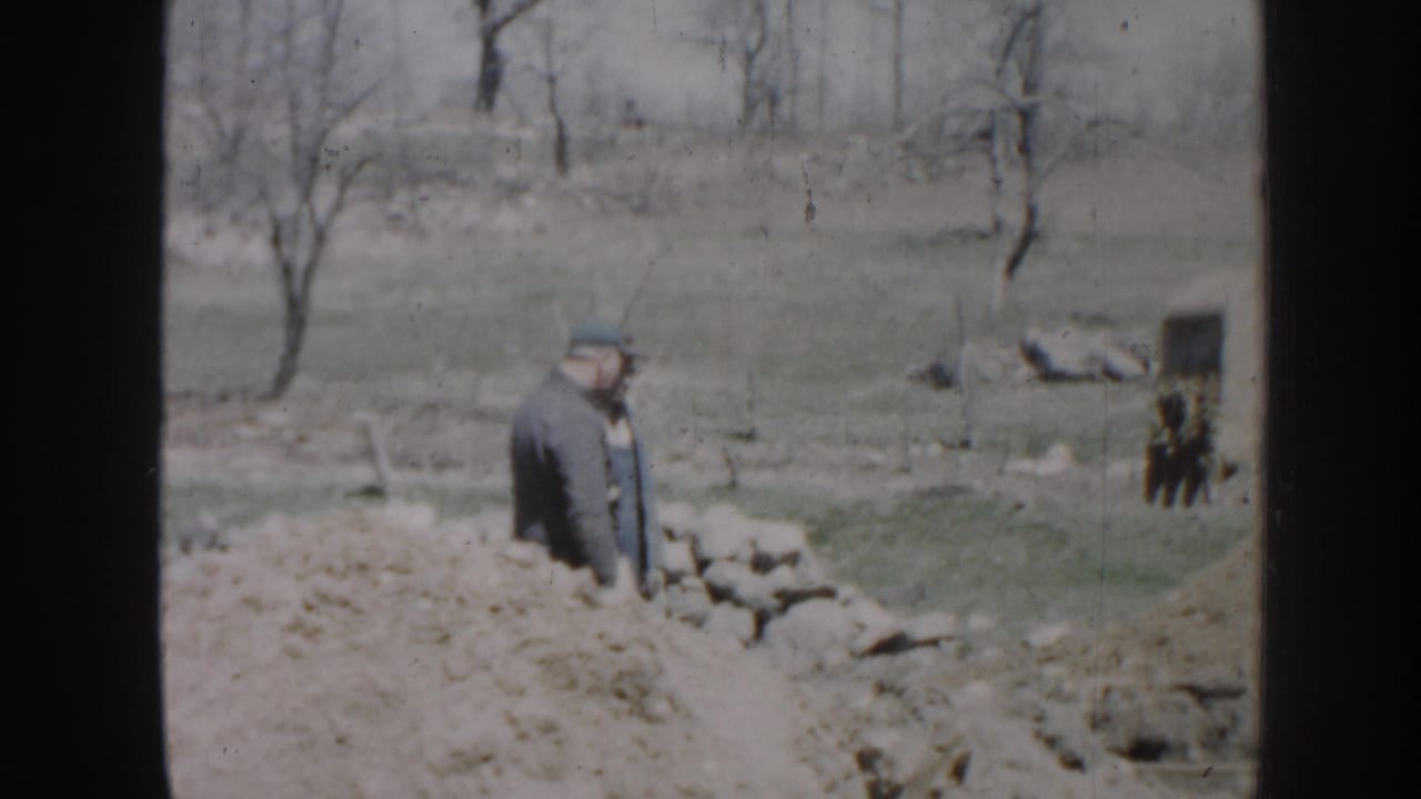 Man in a dark jacket and cap standing near a trench in an open yard.