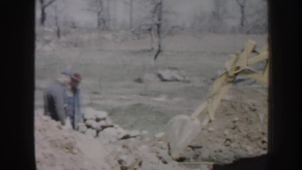Home-movie frame from Nebraska in 1958 showing the backhoe bucket at a dirt trench as a man observes nearby.