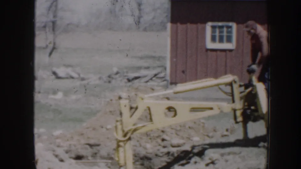 1958 Nebraska home-movie frame showing a yellow backhoe arm, red barn wall, and trench in a rural yard.