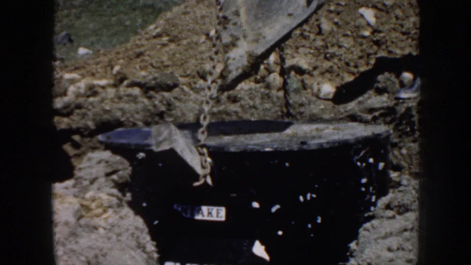1958 Nebraska 8mm frame showing a dark utility vault being lowered into a rural excavation by chain rigging.