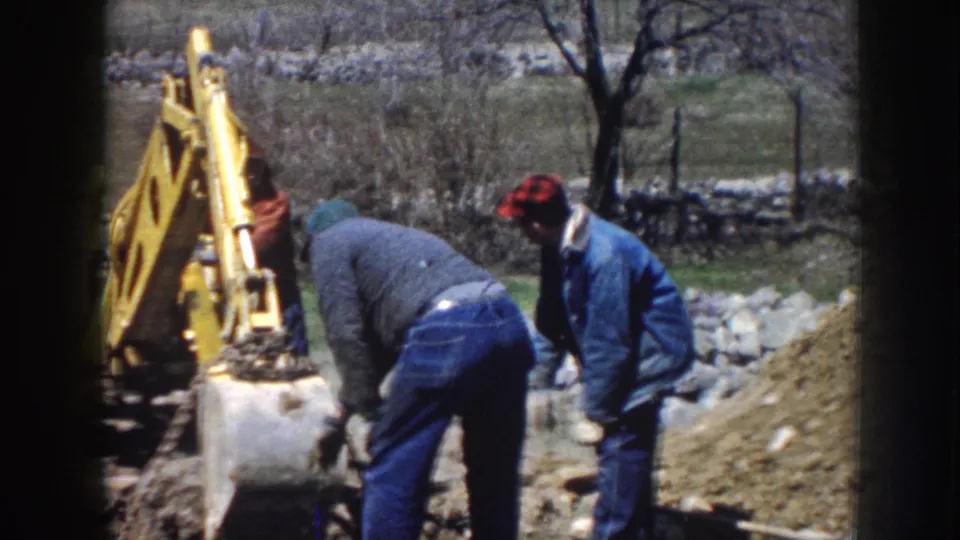 1958 Nebraska 8mm frame showing two workers guiding work beside a yellow backhoe at a rural dig site.