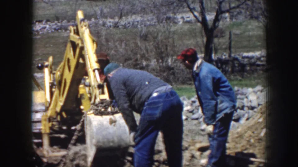 1958 Nebraska construction frame with two workers near a yellow backhoe bucket in a grassy rural work area.