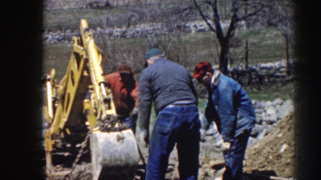 Three men in work clothes stand beside a yellow backhoe bucket near a dirt pile.