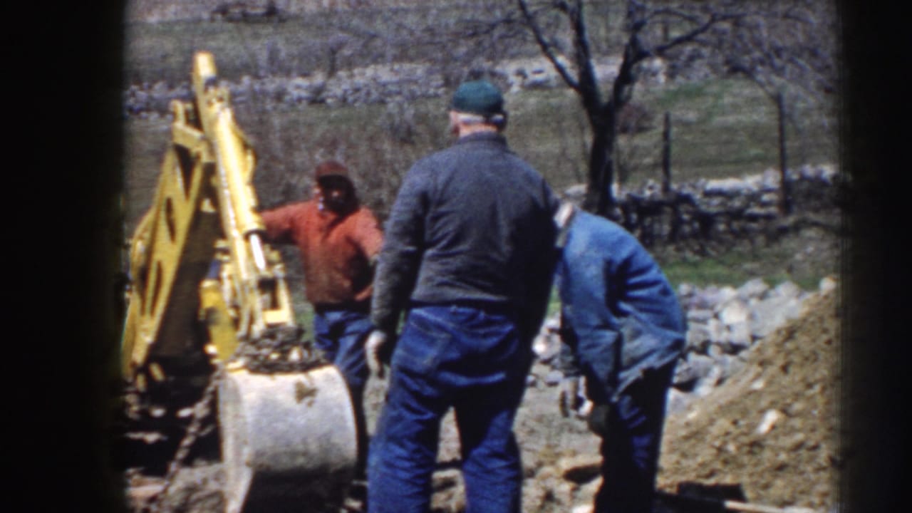 Three men in jackets and caps remain near the yellow backhoe, with rural ground and a stone wall behind them.