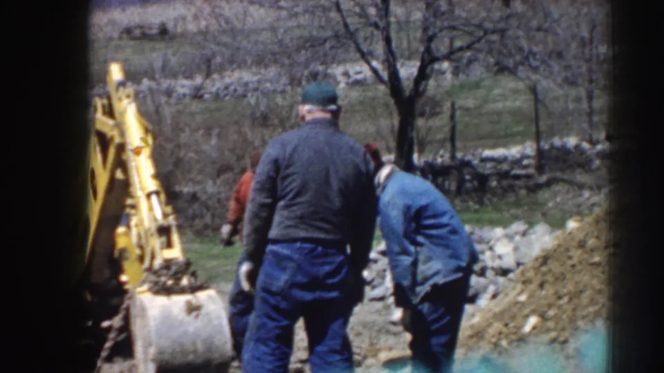A 1958 Nebraska home-movie frame shows men near a yellow backhoe, with one figure turned away and another bent over the work area.