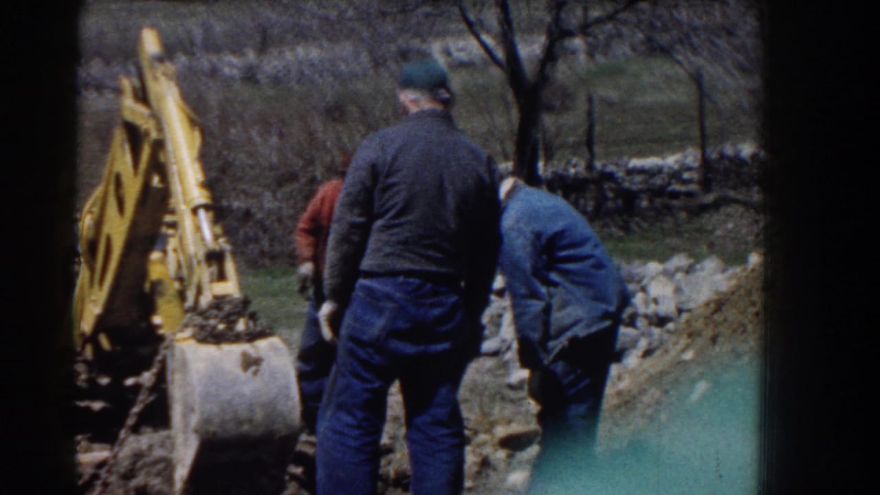 Men stand near a yellow backhoe and dirt area during an outdoor hoisting task.