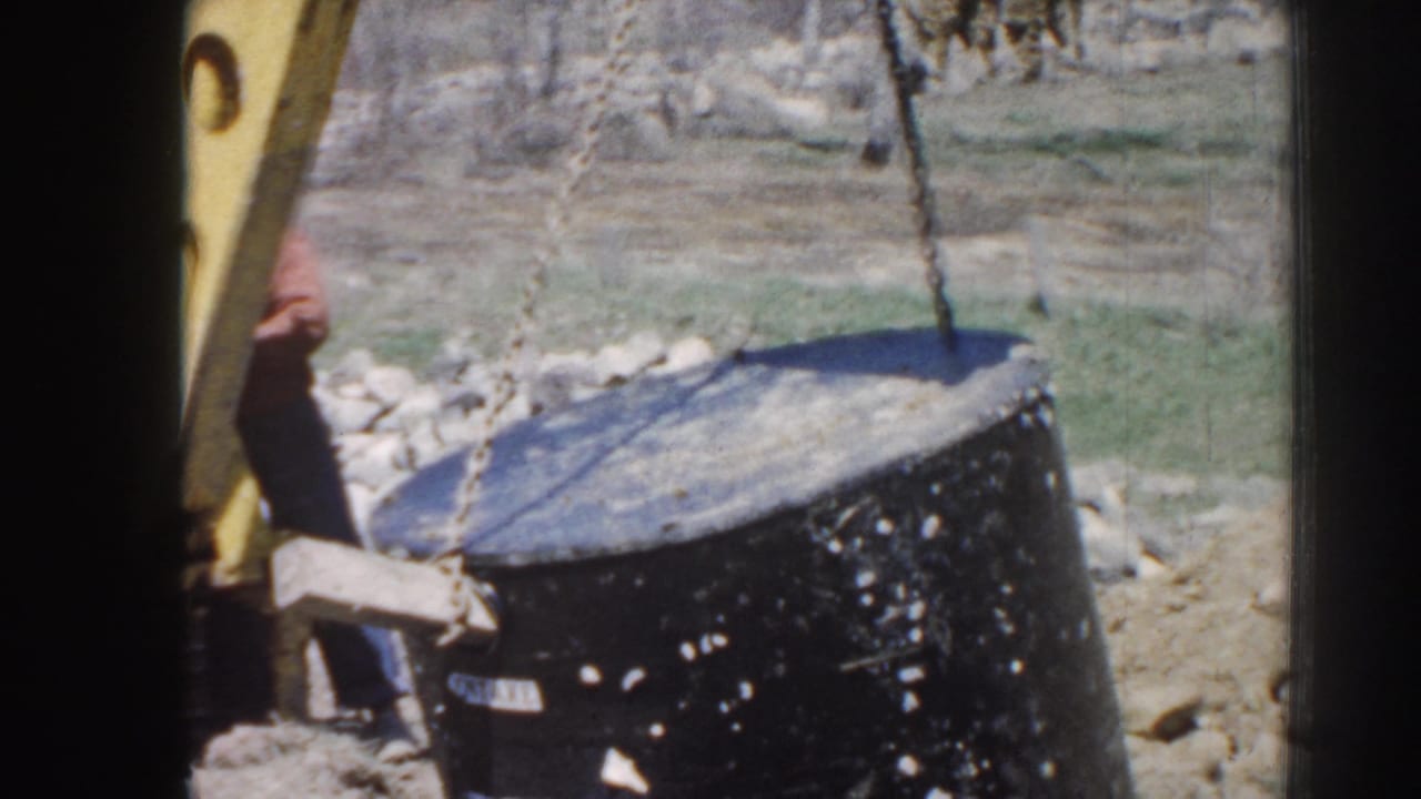 A chain lifts a large black cylindrical object beside a yellow machine arm.