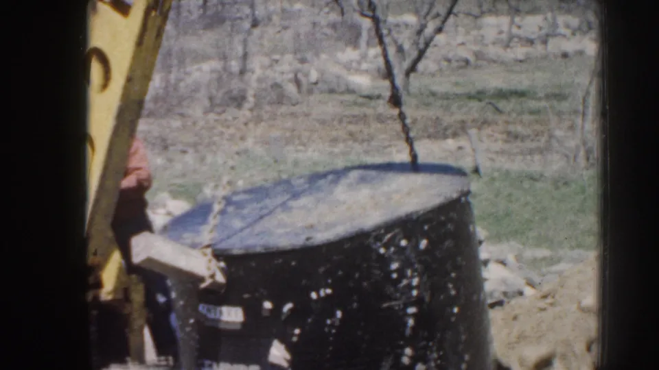 The large black cylindrical object remains suspended by a chain near a yellow backhoe arm on open rural ground in Nebraska.