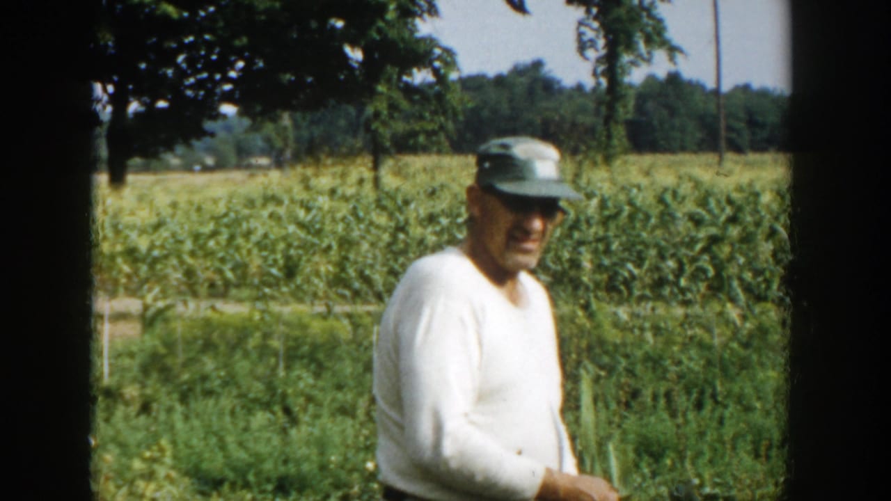 In Michigan 1958 home-movie footage, a farmer in a white shirt and green cap stands near corn rows with trees behind him.