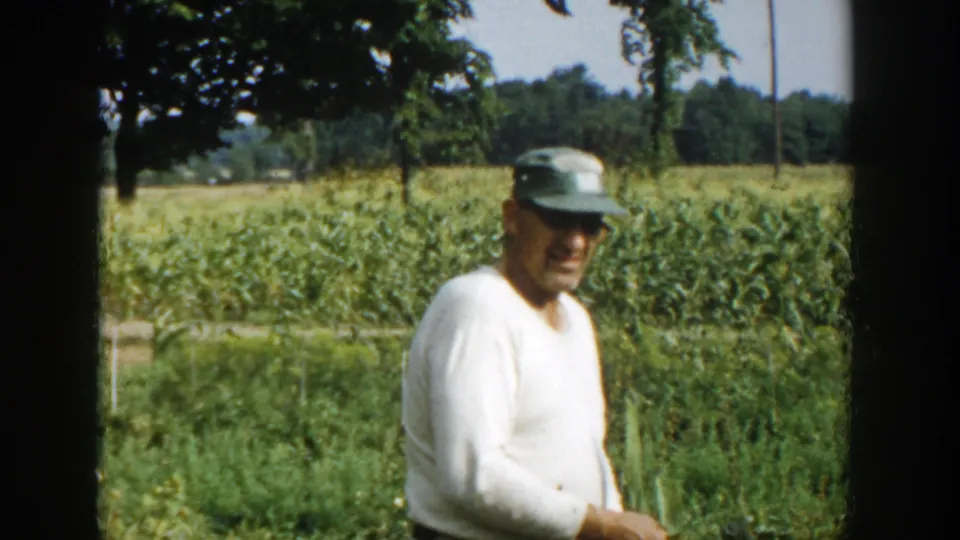 In Michigan 1958 home-movie footage, a farmer in a white shirt and green cap stands near corn rows with trees behind him.