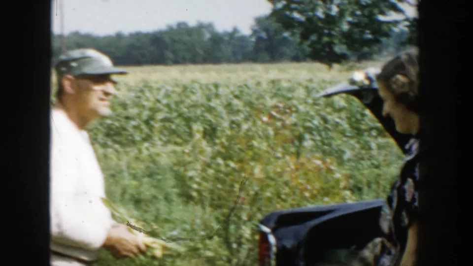A man in a light sweater holds corn while speaking near a woman and the open rear of a parked car by the field.