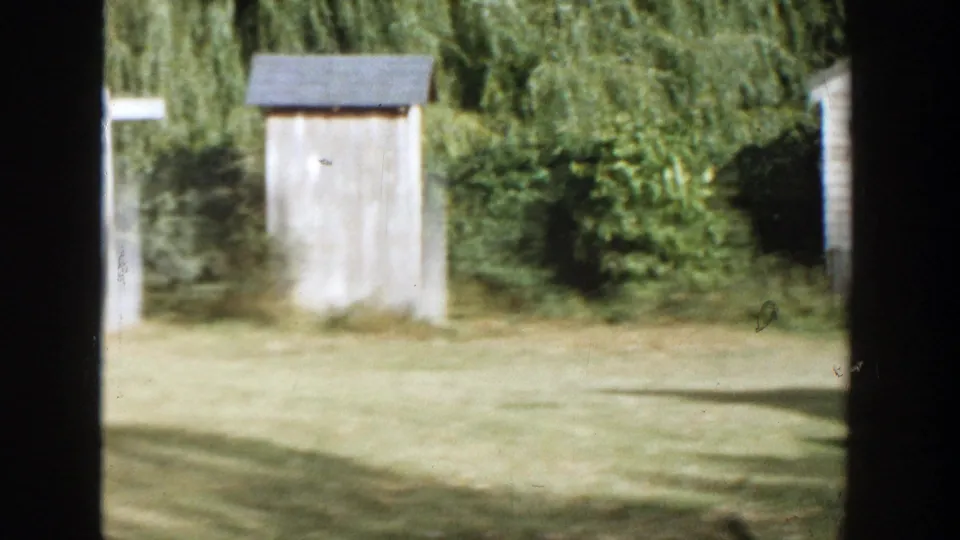 1958 Michigan farmyard view with a small wooden outbuilding, trees, and open grass.