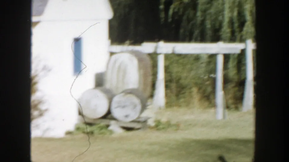 Rural Michigan 8mm frame showing a white gate, barrels, and part of a light-colored building.