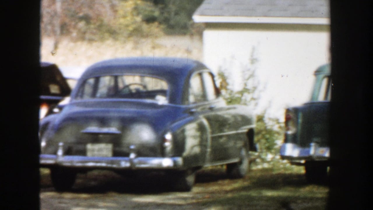 Rounded blue sedan and dark green station wagon parked beside a white garage or house.