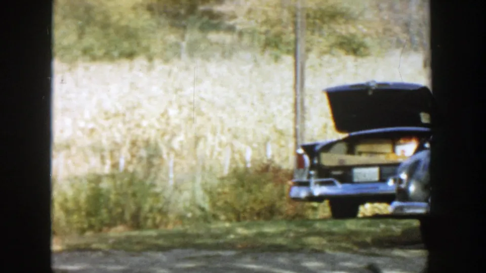1958 Michigan home-movie frame showing a parked car with its trunk open, another car at right, and tall grass behind them.