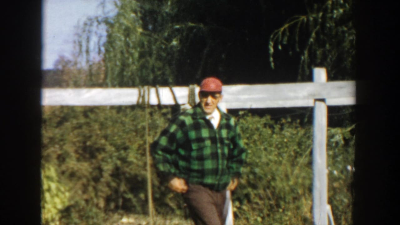 Adult man in red cap and green plaid shirt faces the camera near a white fence in 1958 Michigan footage.